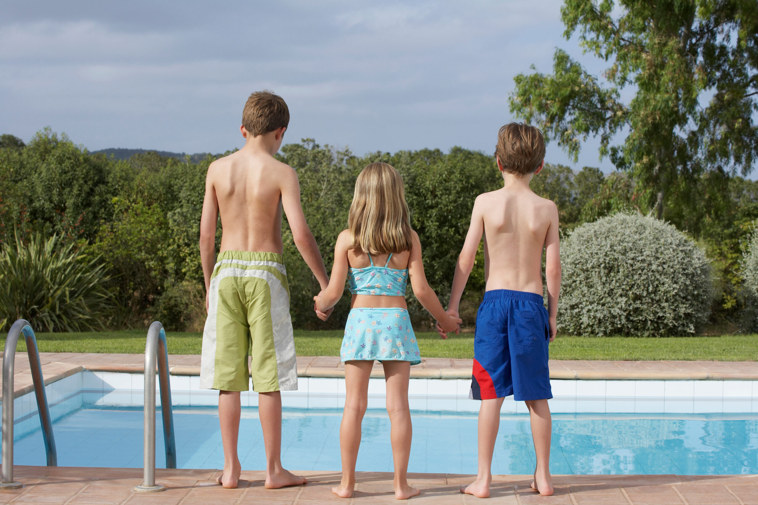 Full length rear view of two brothers and a sister at the edge of an unfenced pool, symbolizing an attractive nuisance.