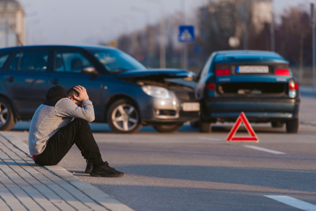 Image of a person sitting on the side of the road holding his head. The scene of a car accident is in the background. He has collided with an uninsured or underinsured driver.