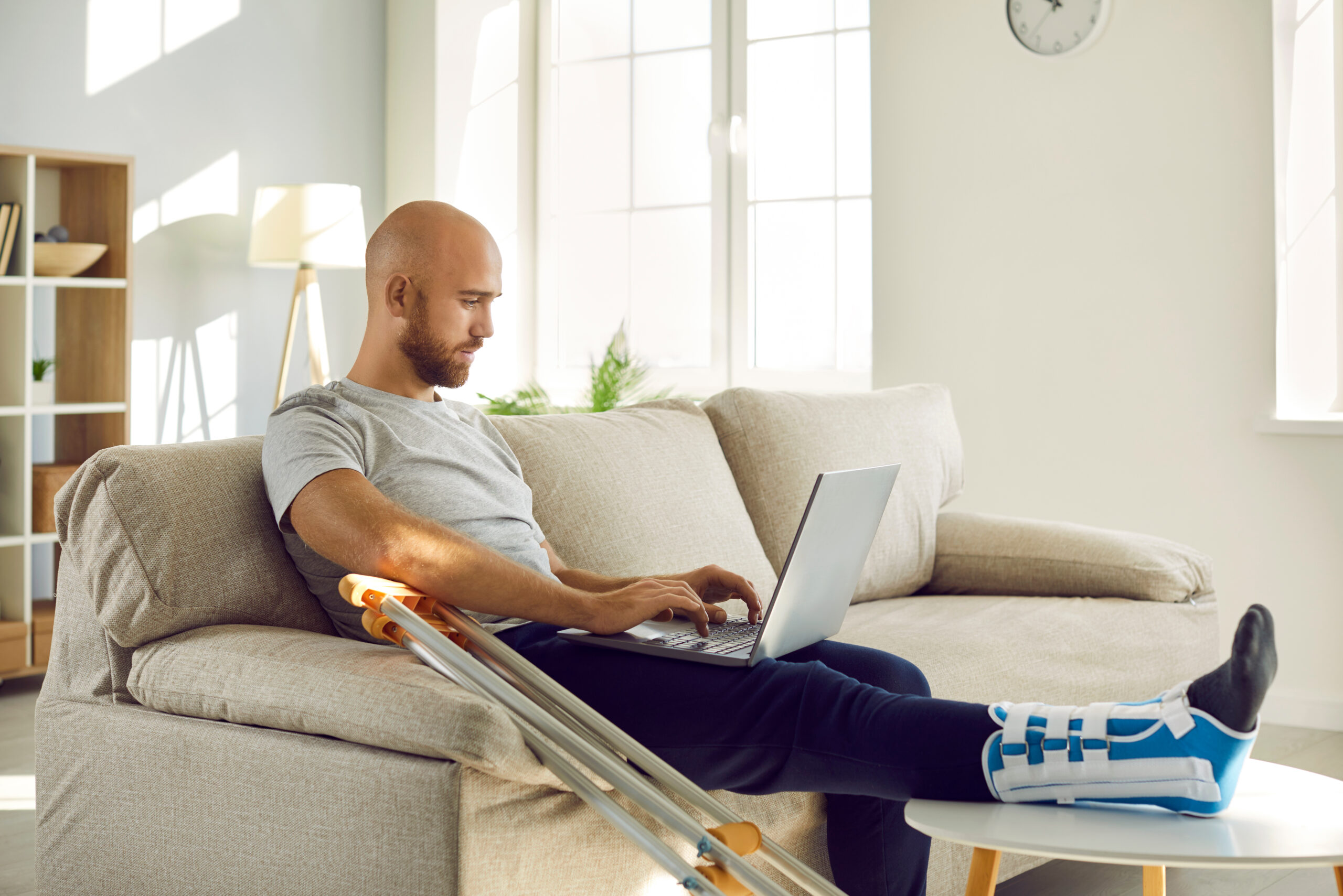 A man has his leg in a cast and crutches beside him as he sits on the couch. He is researching workers