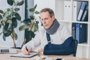 Man in a neck brace and arm sling filling out an injury report for his workers' compensation claim.