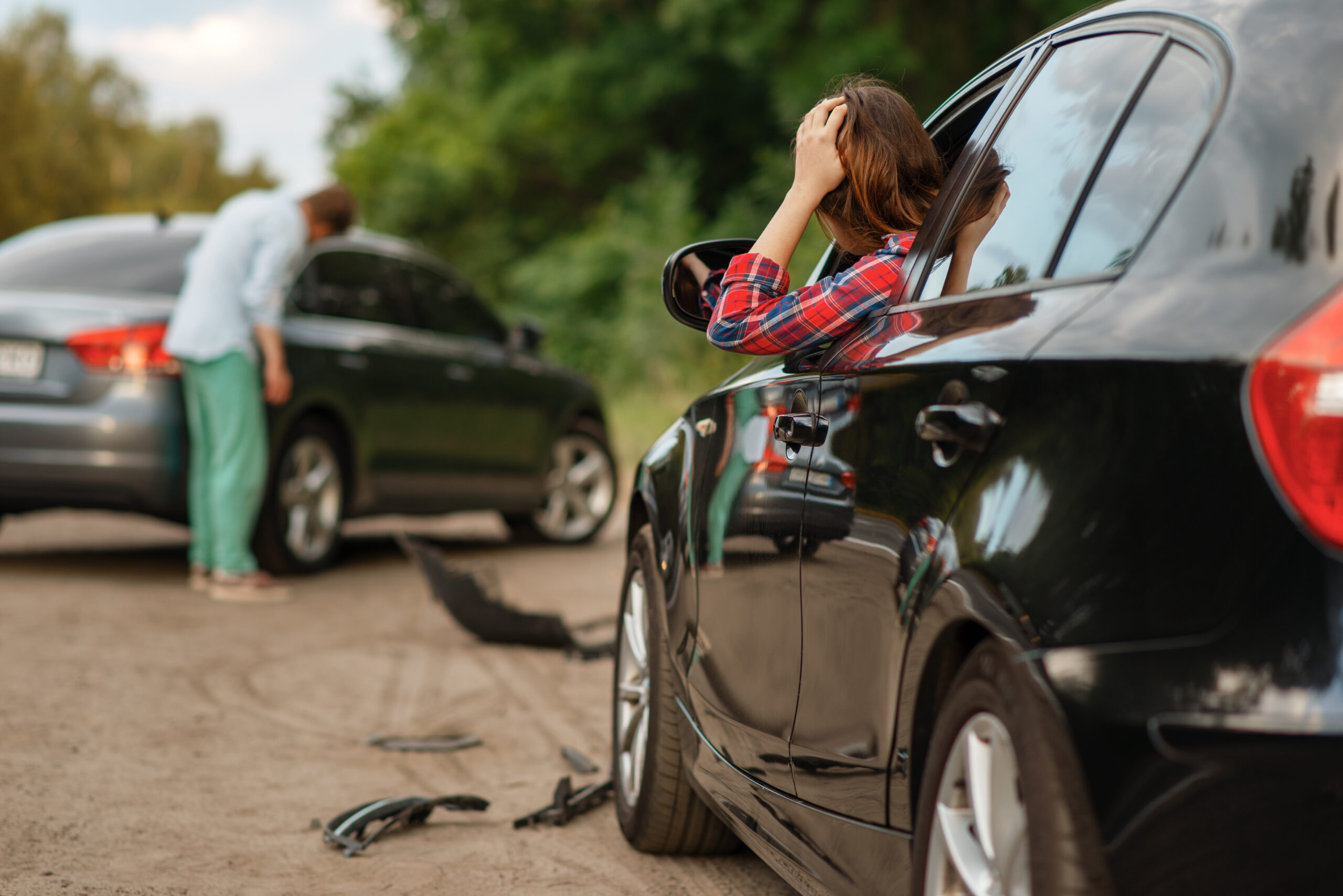 Image of a car accident scene. A woman hangs out of her driver-side window holding her head to look at the damage. A man in the background is surveying the damage to his car