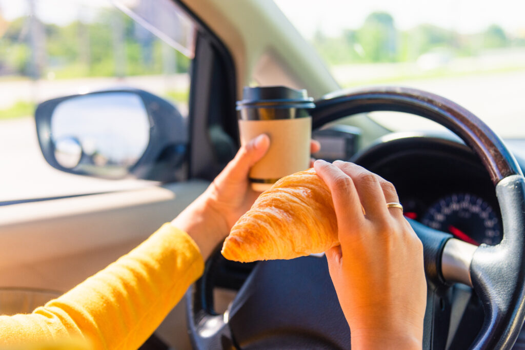 Closeup of a woman's left hand on the steering wheel while holding a coffee. She holds a croissant in her right hand.