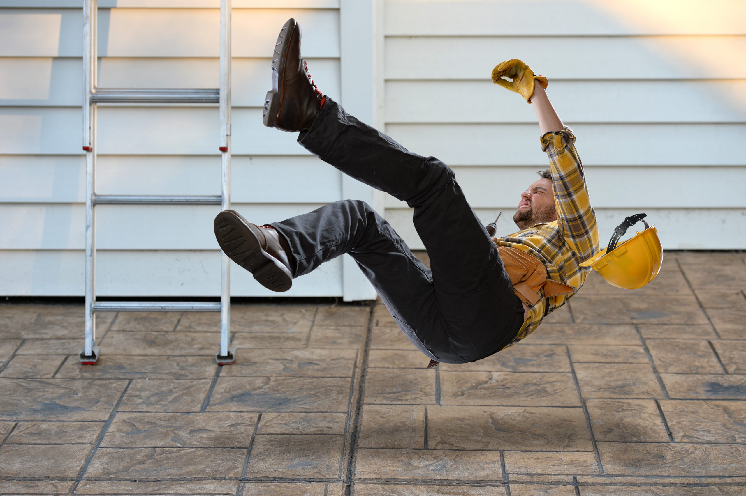 Worker with hard hat falling from a ladder onto a concrete floor. The ladder is propped up on the side of a house.